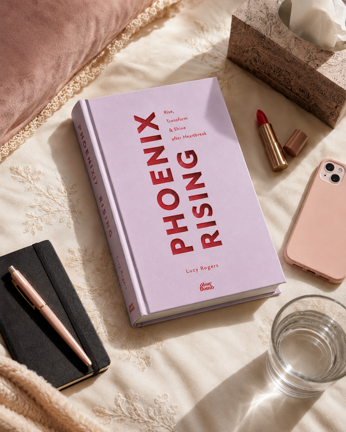 Phoenix Rising by Lucy Rogers, styled on a bed beside a lipstick, notebook and glass of water